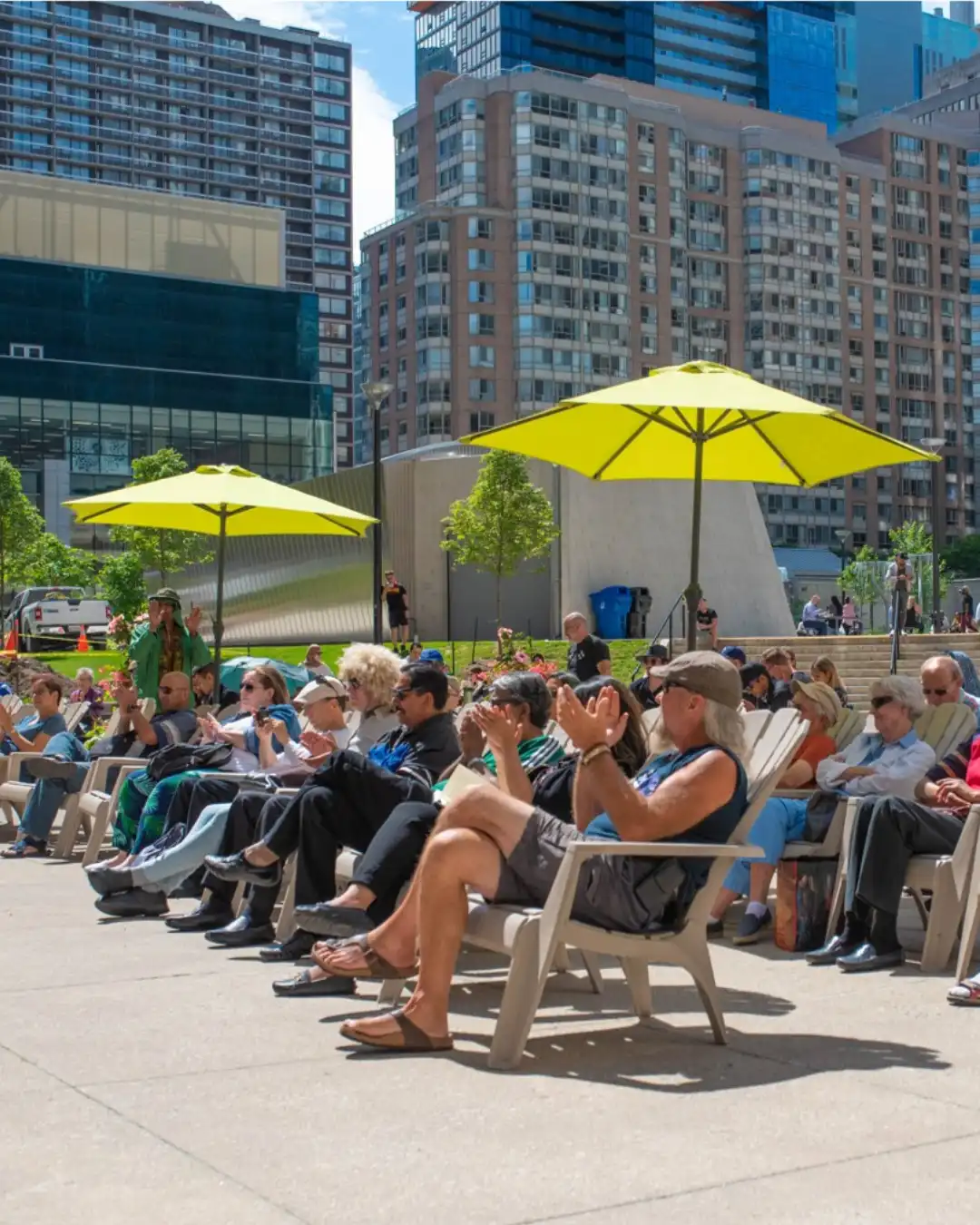 Crowd of people sitting on Muskoka chairs, outside in a park, clapping.